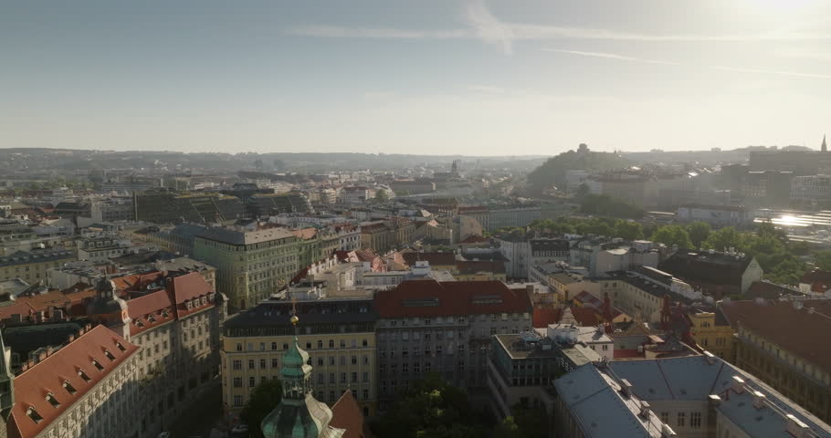 Sunlit Prague cityscape from above with red roofs, surrounding hills and hazy afternoon light.