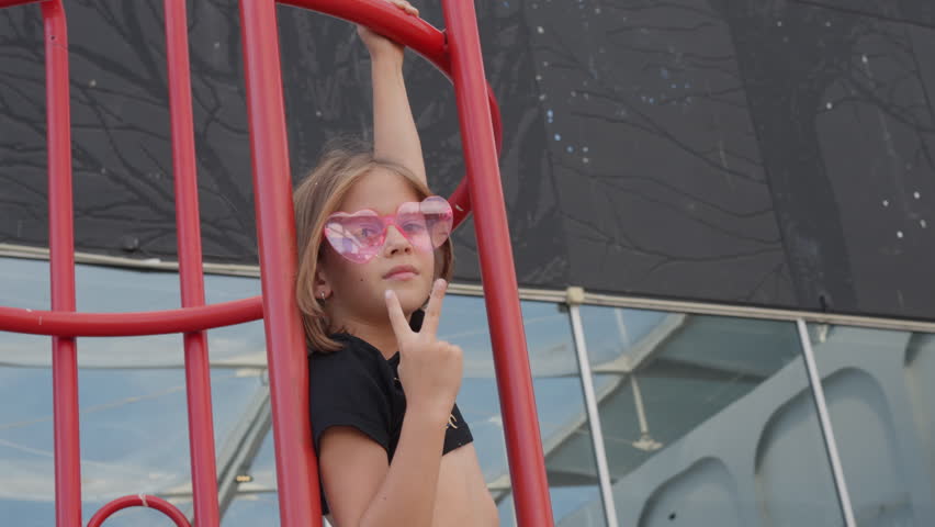 Young girl in stylish heart shape sunglasses hanging on red gate while showing peace sign during daytime