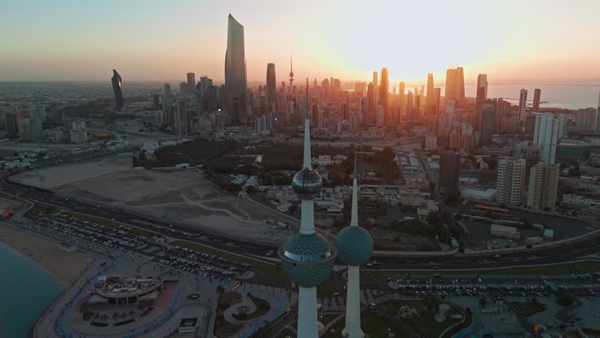 Cinematic drone aerial of Kuwait Towers at golden hour with Kuwait City skyline, seafront roads, and Arabian Gulf horizon glowing under warm sunset light.