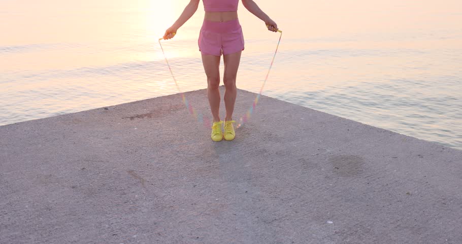Fit young woman jumping rope at the beach against sea and sky background. Summer sport and healthy lifestyle concept. Slow motion