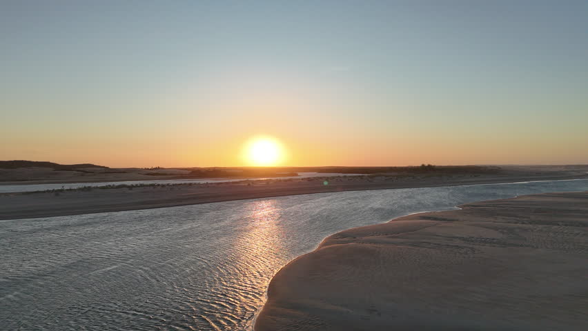 Aerial view of a sunset at the mouth of the Mundaú River in the village of Mundaú, Ceará, Brazil, with warm golden light reflecting over the water and surrounding coastal landscape.