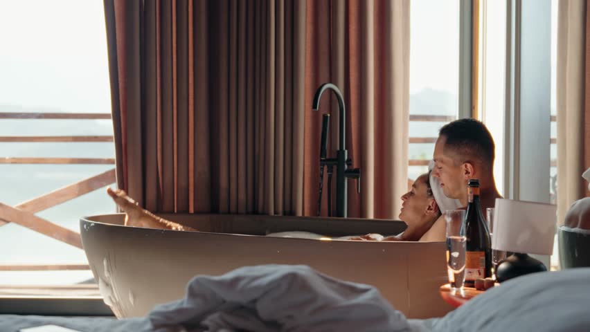 Romantic couple enjoying a hot bubble bath together while clinking champagne glasses in a rustic wooden cabin. Morning light pours in through the window, creating an intimate and serene atmosphere