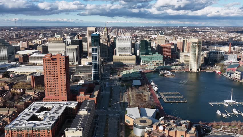 Baltimore downtown skyline with modern skyscrapers, historic buildings and Inner Harbor waterfront under dramatic clouds, showing an iconic American cityscape. Aerial lateral wide shot.
