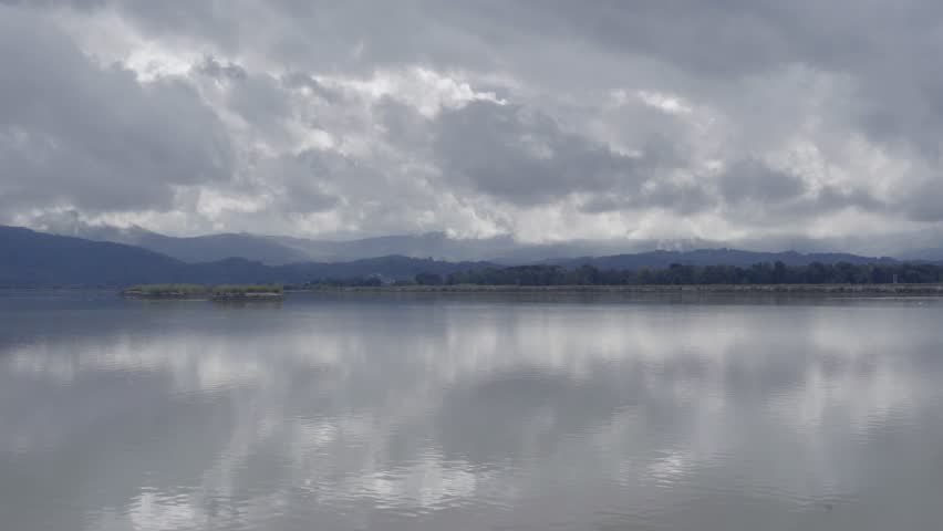 A wide panoramic shot of the Sava River in the Posavje region. The calm water surface perfectly reflects the cloudy sky and distant hills.
