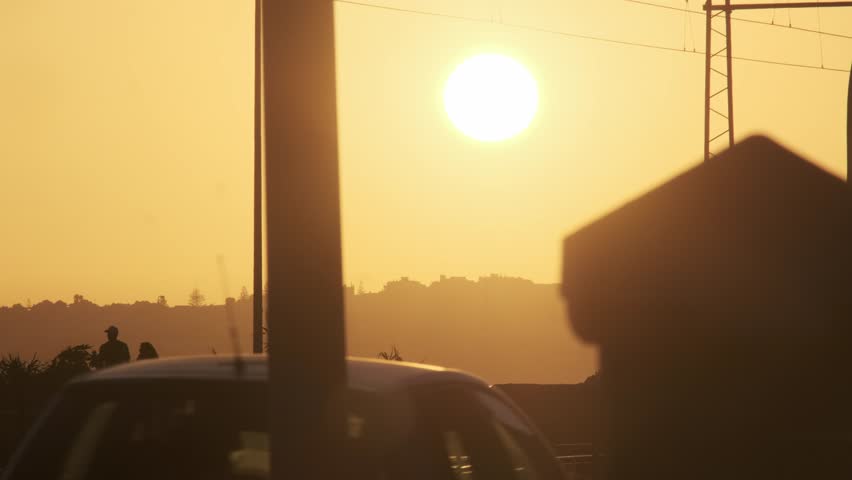 Backlit commuter train moves along electrified tracks during golden hour. Warm sunset light creates dramatic silhouettes of rails and overhead lines.