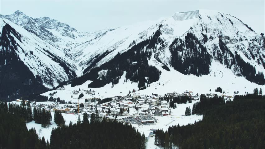 A panoramic winter view of Berwang-Bichlbach ski-resort in Austria: a charming alpine ski resort village nestled in snow-covered mountains, with pine forests, ski slopes, chalets, and winding roads under a cold, overcast sky.