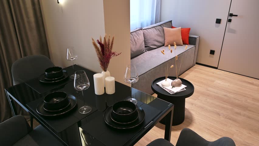 A modern living-dining area with a grey sofa, black dining table set with blackware, round side table, dried floral decor, neutral tones, light wooden flooring — chic and cozy.

