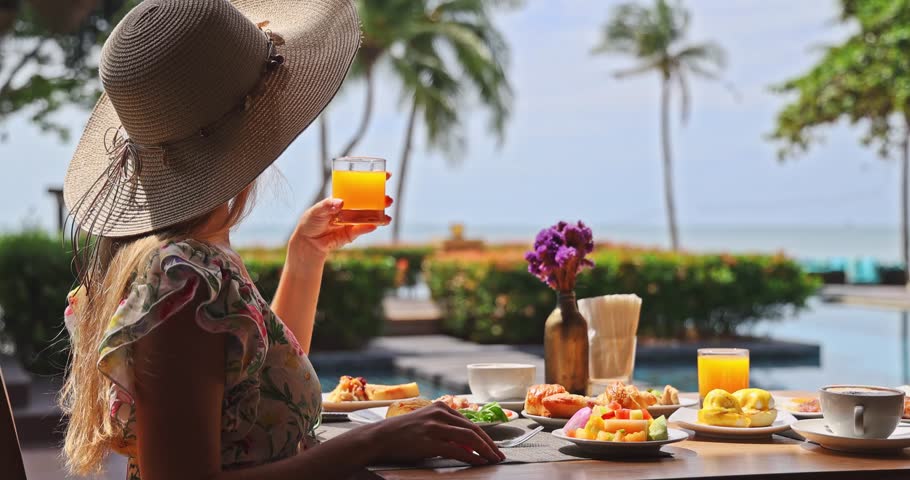 Woman wearing straw hat holding orange juice glass at poolside breakfast table, tropical ocean view with palm trees, bright sunny morning, luxury travel lifestyle, vacation relaxation.