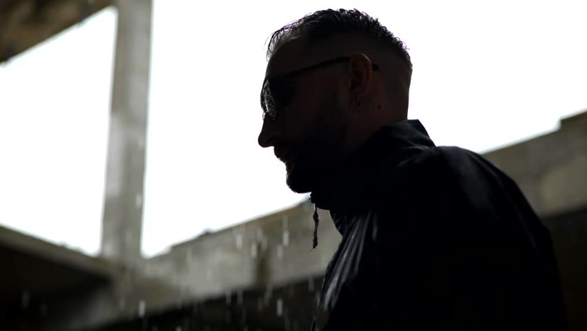 Man in black attire standing in a decaying, damp interior as water streams from the ceiling. Dripping water illuminates a dark, unfinished concrete space where a suited figure stands. Depressed man in the rain.