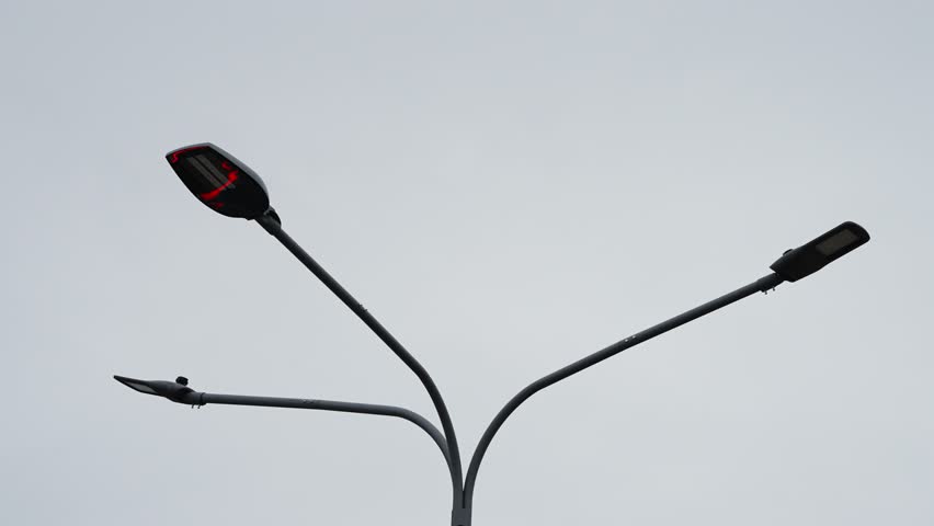 Modern triple-headed streetlight with curved arms rising into an overcast grey sky, capturing minimalist urban infrastructure and symmetrical design along a quiet roadway.