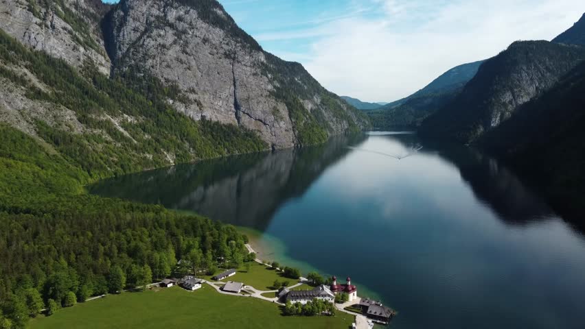 Breathtaking aerial view of the famous Königssee lake in Bavaria, Germany. The shot captures the emerald water, steep fjord-like mountains, and the historic St. Bartholomä church on the peninsula.
