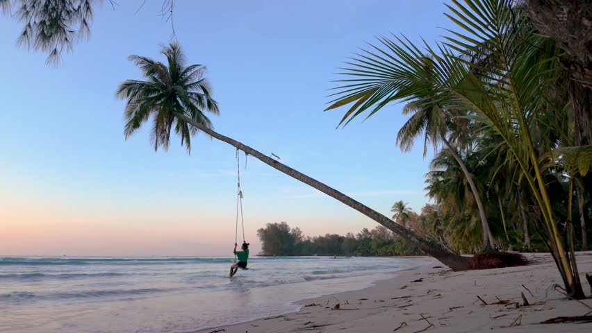 Gentle waves lap at the shore as a woman swings from a palm tree in Koh Kood Thailand. The sun sets, casting a warm glow over this tropical paradise. A perfect spot for relaxation and adventure.