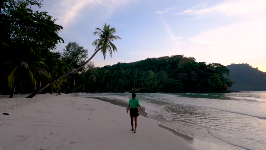 A lone woman traveler strolls along the pristine shores of Koh Kood, Thailand, as the sun sets in the background, casting a warm glow over the tranquil landscape and palm trees.