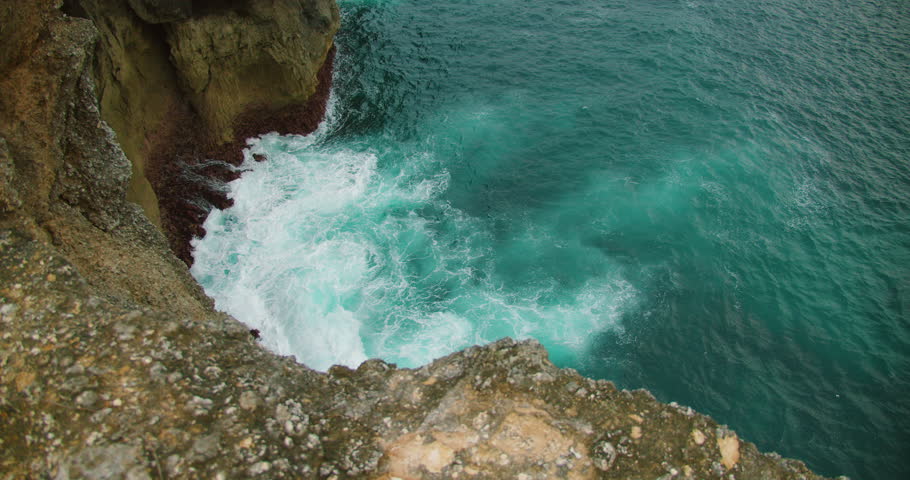 Top down view of turquoise water violently crashing against jagged dark coastal rock formation
