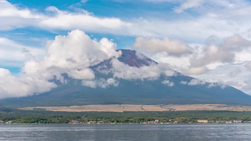 Mount Fuji with clouds, green trees, calm water and clear skies on horizon. Serene scenery of Japanese natural landmark and landscape with mountain partly covered by clouds