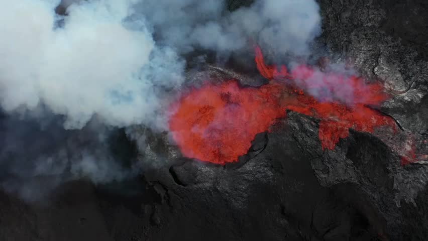 Drone captures a top-down shot of active Kilauea volcano. Molten lava flows, contrasting with black volcanic rock and white smoke rising. Hawaii Volcanoes National Park.