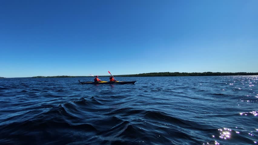 Two people enjoying a sunny day of kayaking, paddling together in a tandem kayak on the beautiful wavy blue water of a large lake with a clear sky and distant shoreline on the horizon
