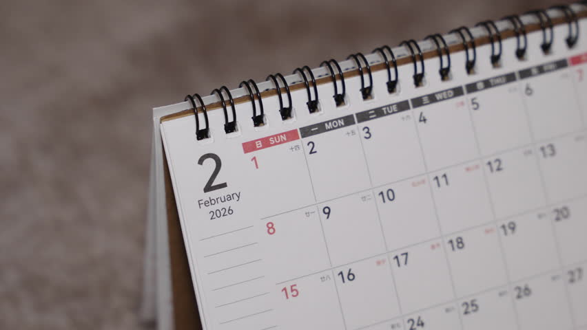 Close-up of a desk calendar page being flipped from February 2026 to March 2026. Hand turns the paper forward while the camera remains static, showing a smooth monthly time transition indoors.