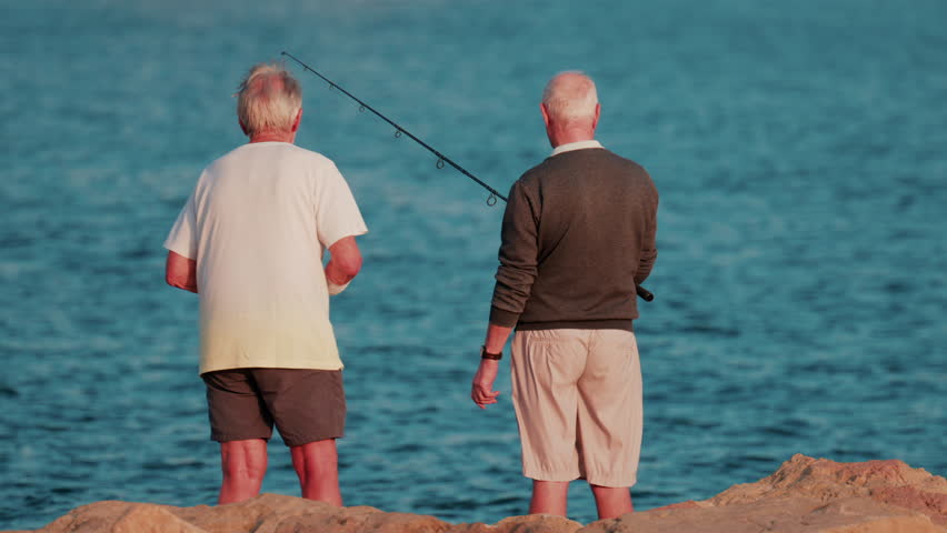 Rear view of two elderly men standing side by side on the rocks, looking at the horizon while fishing