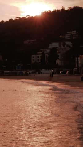 The sun sets over a coastal town with trees, hills, and buildings reflected on the golden sea. Vertical