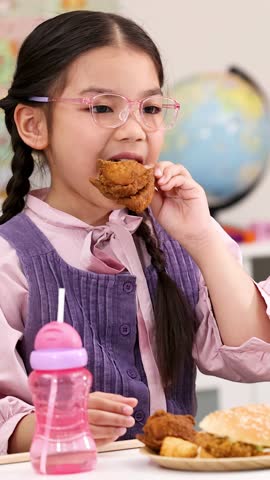 Young girl with braids enjoys fried chicken meal at school desk, bright classroom lighting