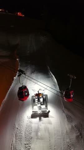 Night aerial drone view in Ortisei, Dolomites. Snow grooming tractor climbs mountain slope compacting snow and shaping fresh ski pistes, while red cable car cabins glow above, preparing resort for morning skiing