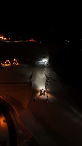 Night aerial drone view in Ortisei, Dolomites, Italy. Snow grooming tractor climbing mountain slope, compacting snow and preparing fresh ski pistes for morning alpine skiing under dark winter sky
