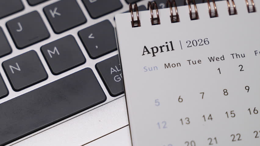 Close-up of a desk calendar with April 2026 visible on a laptop keyboard. Slow rightward camera rotation presents clear date structure and structured monthly planning concept indoors.