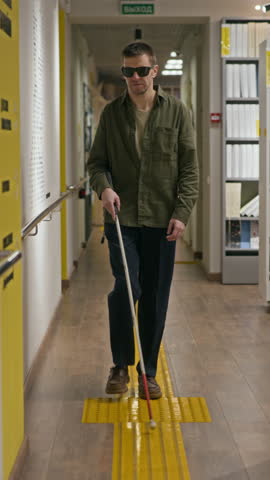 Vertical slowmo shot of middle-aged Caucasian man in dark glasses using walking stick while moving around modern library with yellow tactile indicator for blind and visually impaired people