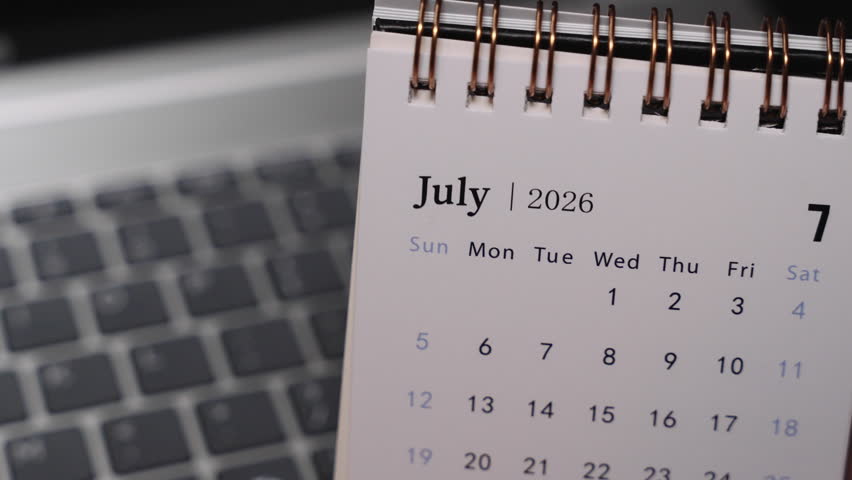 Close-up of a desk calendar on a laptop keyboard as a hand flips the page from July 2026 to August 2026. Camera stays static, presenting a clear monthly time transition and organized planning concept indoors.
