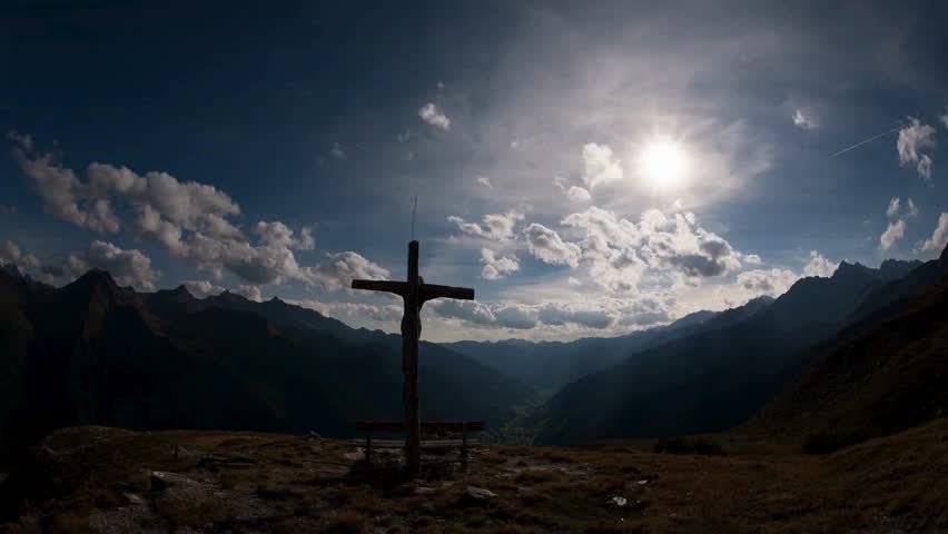 Wooden Christian cross stands atop a mountain peak overlooking a valley. Dramatic clouds fill a blue sky with sun shining down. Religion, nature, faith.
