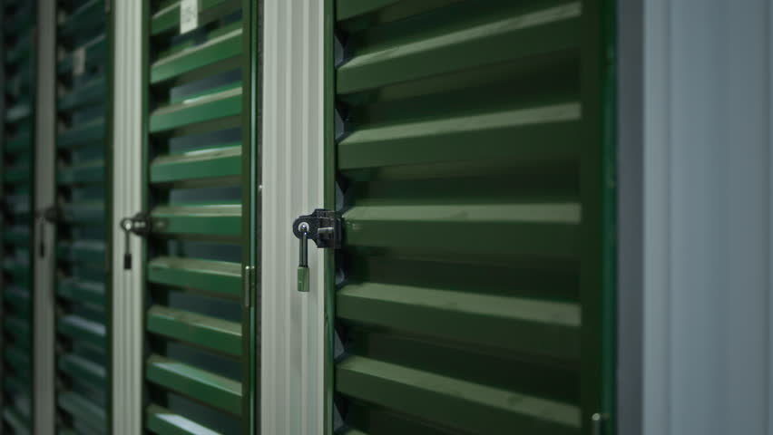 Row of green metal storage unit doors in corridor of self storage facility, each secured with padlocks