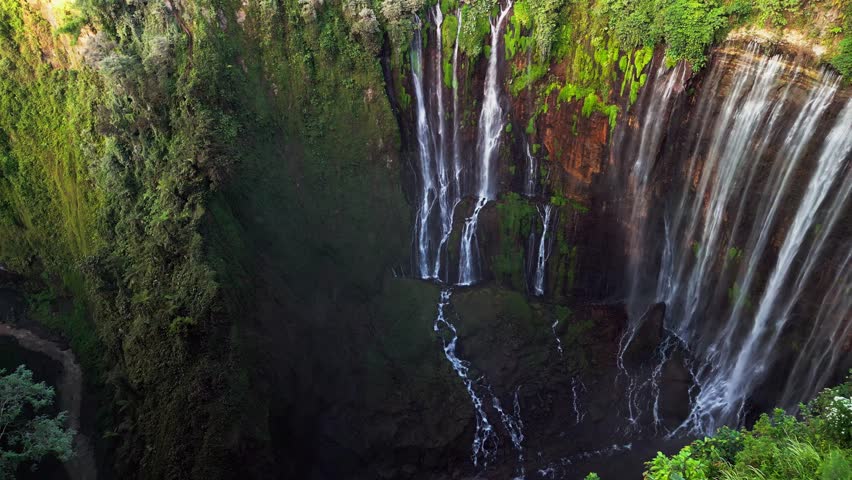Aerial footage showing the cascading waterfalls of Tumpak Sewu in Indonesia as multiple streams descend into a deep limestone gorge with visible river flow drifting mist and rugged tropical terrain