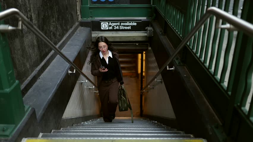 Professional young businesswoman with glasses talking on her smartphone while walking up the stairs and exiting the 34th street penn station subway in new york city, carrying a handbag