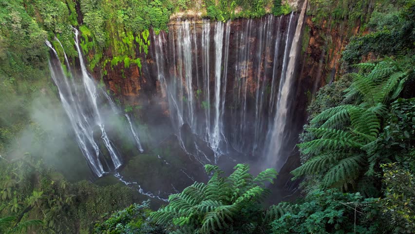 High angle drone video capturing Tumpak Sewu Waterfall in Indonesia with countless water streams flowing over steep canyon walls creating heavy mist, lush greenery and a powerful tropical landscape.