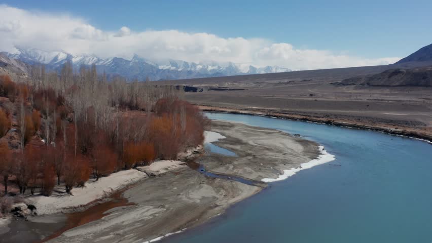 Aerial view of turquoise Indus River flowing through winter Ladakh valley lined by brown reed banks under cloudy skies with distant snowy peaks.
