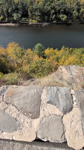 Vertical video featuring a slow pan up from stone overlook to a calm river and dense autumn forest in the Catskills. Colorful fall foliage lines the water with rolling hills in the background.