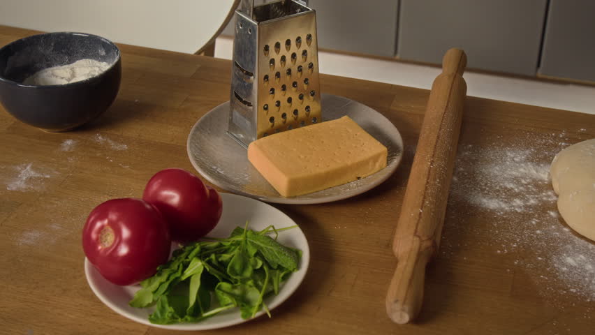Close-up shot of flour, tomatoes and fresh greens, chunk of cheese and grater, wooden rolling pin, dough piece, sliced pepperoni and small bowl with red sauce on kitchen table