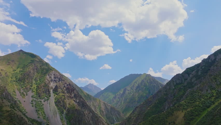Vast mountains rise majestically with green slopes and rocky outcrops. Serene valley below contrasts with blue sky and fluffy clouds. Ascending drone view.
