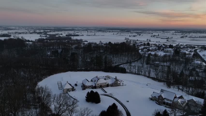 Aerial view of large countryside mansion on rolling hills at sunrise, surrounded by snowy fields and trees. Peaceful American winter landscape from above. Wide shot. Snowy winter dusk.