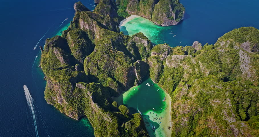 Aerial flight over Phi Phi Leh island revealing limestone cliffs, lush green vegetation, and clear turquoise water of Maya Bay, with boats navigating around the pristine tropical paradise in Thailand