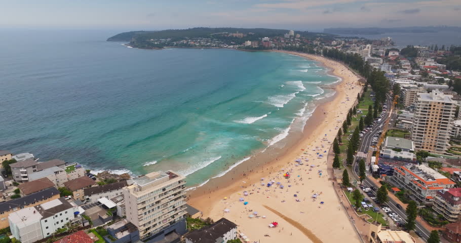 Aerial view of Manly Beach in Sydney, Australia, people relaxing on sand, swimming in ocean waves, and buildings lining the coastal road on a clear day. Drone flight over shoreline cityscape landscape