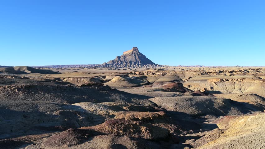 A wide landscape drone shot captures Factory Butte, a prominent geological feature in Utah. The isolated butte stands above a vast, barren desert and grey badlands terrain under a clear blue sky.