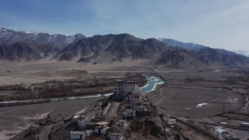 Winter aerial captures Stakna Monastery atop rocky cliff overlooking Indus River, barren valley and distant snow peaks in Ladakh.