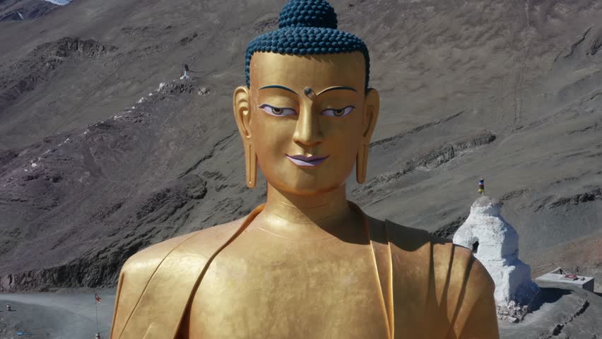 Drone pull out from detailed golden Buddha statue widens to reveal stupa, buildings, valley village and snow-capped Ladakh mountains.