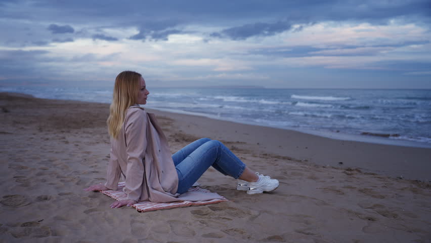 Middle-aged woman in trench coat and jeans sit on sand thoughtfully by moody sea, pensively watch ocean waves crash on shore.Concept mental health,self reflection or therapy visuals,with overlay space