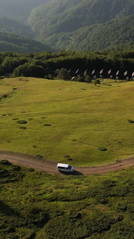 Aerial view of a white van driving along a winding dirt road through lush green hills. A beautiful mountain landscape with dense forests in the background