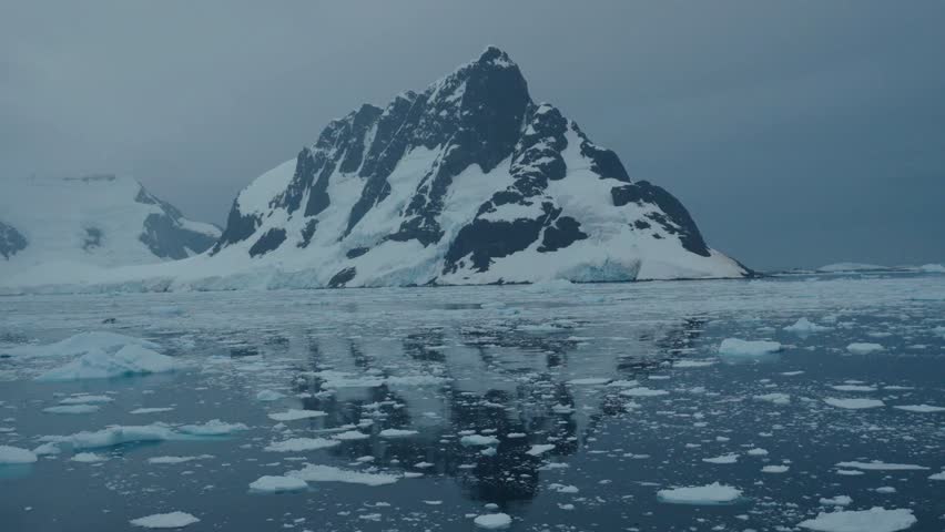 A serene aerial view captures imposing, snow-covered mountains rising from a calm Arctic sea filled with floating ice floes under a hazy sky.