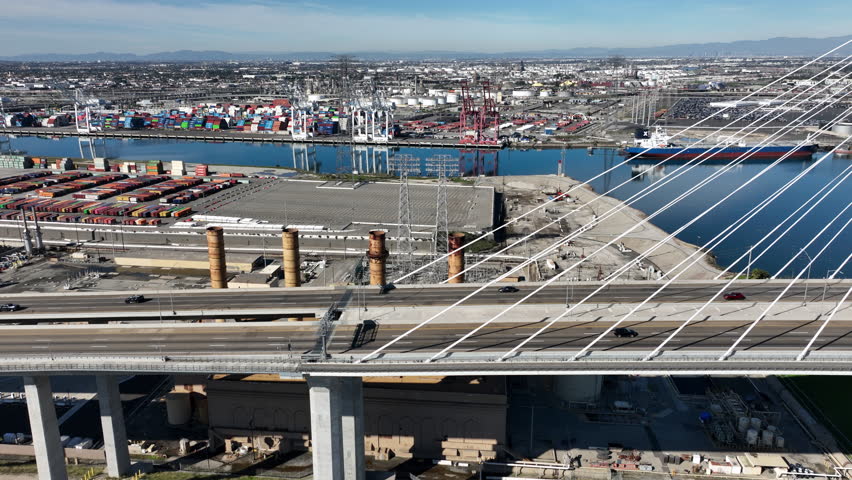 Aerial view of the International Gateway Bridge in Long Beach, California next to Terminal Island, with shipping containers, 18-wheelers, and the Port of Los Angeles visible.