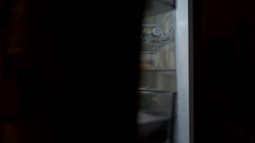 Back view of man drinking yogurt from small bottle while standing in front of open refrigerator in night kitchen. Concept of night eating, unhealthy habits and disrupted diet routine